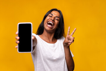 Happy, excited young indian woman showing phone screen to camera doing peace sign gesture with hand.
