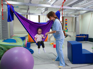 Physical therapist working with little girl in hammock made of elastic material. sensory integration room. Physiotherapy and neuro corrections in children therapy center