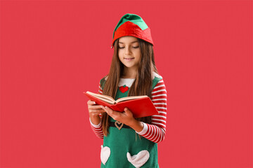 Little girl in elf pajamas reading book on red background