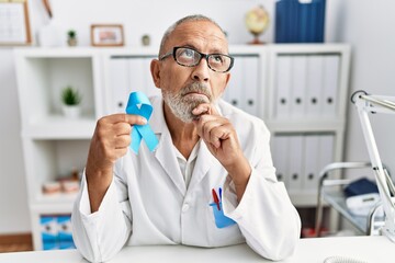 Mature doctor man holding blue ribbon at the clinic serious face thinking about question with hand...