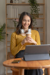 Pretty Asian woman sitting happily working with laptop and drinking coffee smiling and laughing brightly on a comfortable day at home.