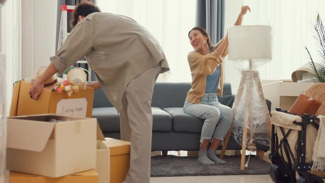 Young happy Asian couple help each other moving box full of stuff unpacking furniture on sofa couch in living room at new house. Husband and wife move new house, Apartment Rent And Ownership Concept.