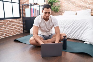 Young hispanic man using laptop sitting on yoga mat at bedroom