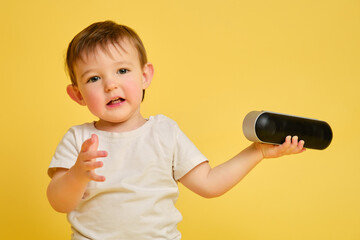 Toddler baby plays with a wireless music speaker on a studio yellow background. Happy child in a...