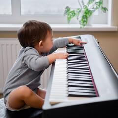 Toddler baby plays piano in home living room, music lessons. Happy child boy learns to play music...