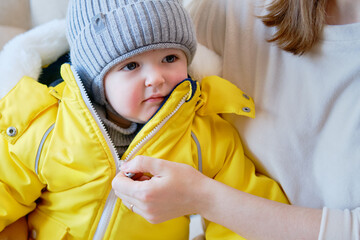Child is dressed in warm winter clothes for a walk outside. Woman mother puts clothes on toddler...