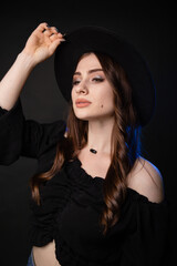 Beautiful young woman in black clothes with a black hat. Photo in the studio on a dark background with blue backlight.