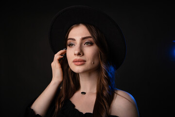 Beautiful young woman in black clothes with a black hat. Photo in the studio on a dark background with blue backlight.