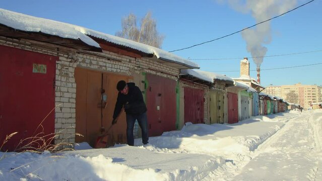 A dexterous man clears snow from the passage to the garage with a wooden shovel. Large snowdrifts around after a snowstorm, sunny day. City life, heavy snowfall. UHD 4K.