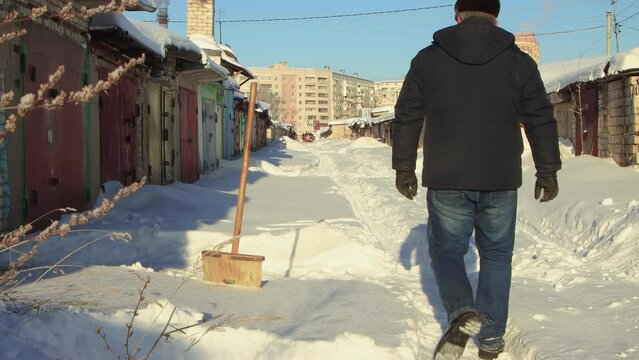 A man cheerfully clears the path to the garage from snow with a wooden shovel. Large snowdrifts around after a snowstorm, sunny day. City life, heavy snowfall. UHD 4K.