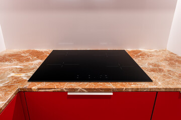 Black induction hob with four burners is built into beige marble countertop of a red kitchen cabinet. Light brown abstract patterned worktop with built-in ceramic hob. Kitchen appliances for cooking.