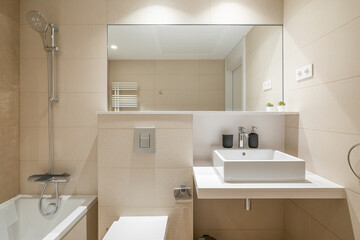 Fototapeta na wymiar Cozy bathroom in modern design with huge mirror on the wall and bright lighting. Walls are in beige tiles and white sanitary ware. Room for taking morning shower and relaxing evening baths.
