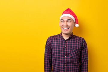 Happy young Asian man in Santa hat looking upwards with broad smile on yellow studio background. Happy New Year 2023 celebration merry holiday concept