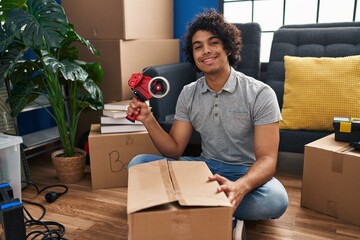 Obraz na płótnie Canvas Hispanic man with curly hair moving to a new home closing cardboard box with tape looking positive and happy standing and smiling with a confident smile showing teeth