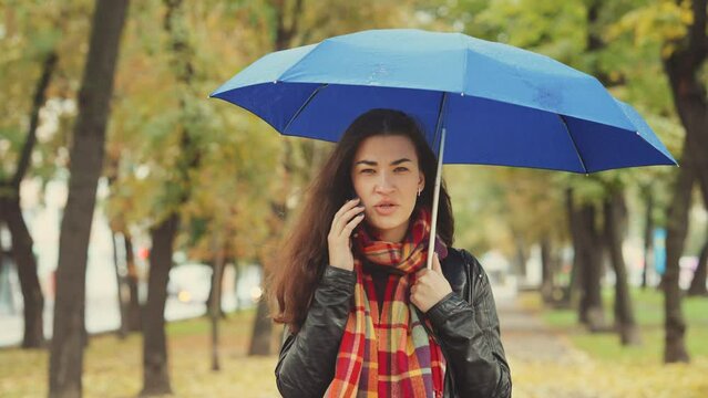 Young woman wearing checkered scarf standing in autumn park under an umbrella during the rain and talking on phone, blurred trees on background