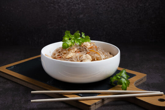 Fried funchosa with chicken in a white bowl with chopsticks on a serving board