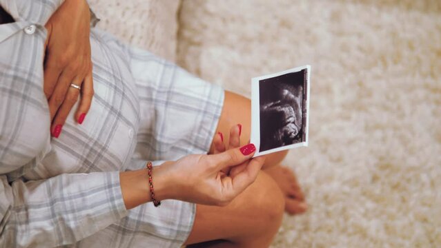 Caucasian pregnant woman sitting on bed, touching belly and looking on ultrasound photo. pregnancy, expecting baby and spending time at home.
