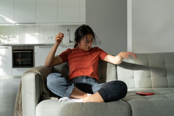 Angry frustrated Asian girl sits on sofa at home throwing away smartphone, getting bad news on cellphone. Young heartbroken woman receiving breakup text from boyfriend. Emotional pain of breaking up