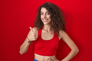 Hispanic woman with curly hair standing over red background doing happy thumbs up gesture with hand. approving expression looking at the camera showing success.