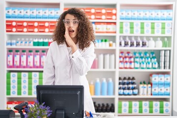 Hispanic woman with curly hair working at pharmacy drugstore hand on mouth telling secret rumor,...