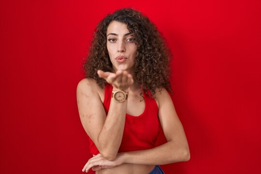 Hispanic woman with curly hair standing over red background looking at the camera blowing a kiss with hand on air being lovely and sexy. love expression.