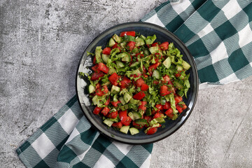 Vegetarian cucumber salad with lettuce, tomatoes and herbs on a round plate on a dark gray background. Top view, flat lay