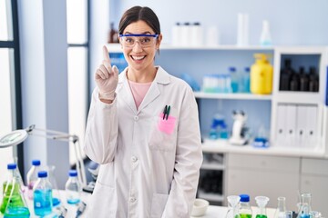 Young brunette woman working at scientist laboratory smiling with an idea or question pointing...