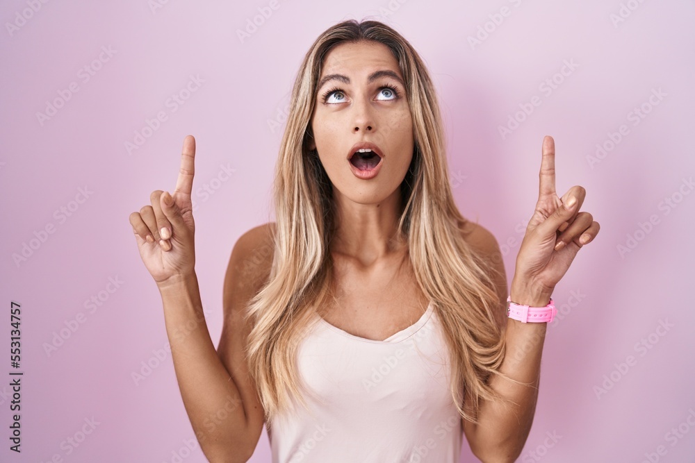 Wall mural young blonde woman standing over pink background amazed and surprised looking up and pointing with f