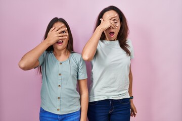 Young mother and daughter standing over pink background peeking in shock covering face and eyes...