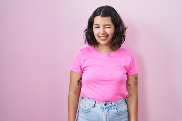 Young hispanic woman standing over pink background winking looking at the camera with sexy expression, cheerful and happy face.