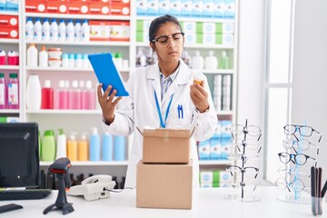 Young beautiful hispanic woman pharmacist using touchpad holding pills bottle at pharmacy
