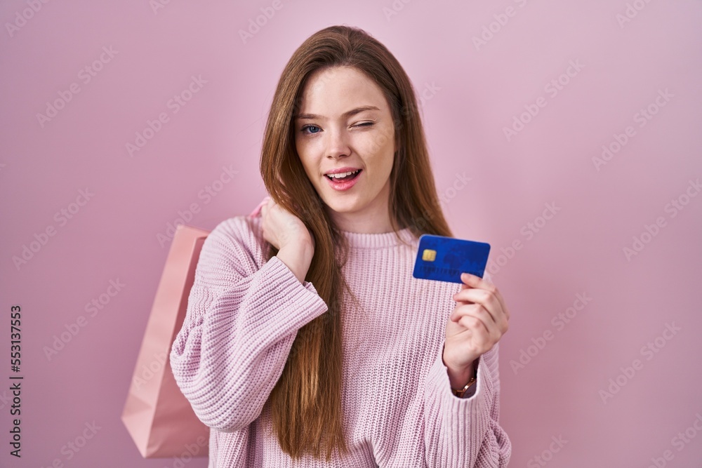 Poster young caucasian woman holding shopping bag and credit card winking looking at the camera with sexy e