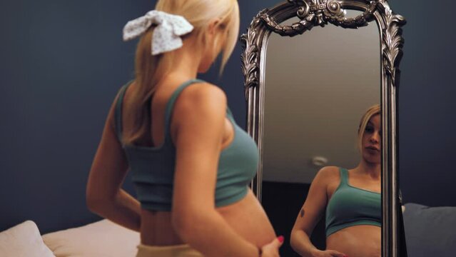 Pregnancy and body care. Attractive blond caucasian pregnant lady in underwear stroking her big belly, doing skin lifting massage, looking at mirror. Mirror reflection