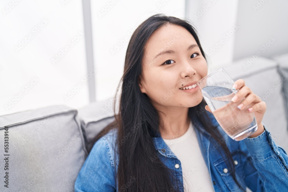 Sticker young chinese woman drinking glass of water sitting on sofa at home