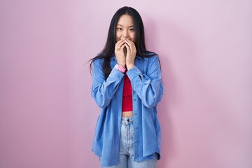 Young asian woman standing over pink background laughing and embarrassed giggle covering mouth with...