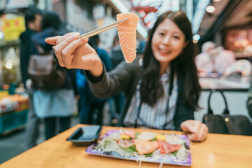 selective focus of a piece of fresh pink Japanese food raw fish sashimi held on chopsticks by an asian female diner at table in kuromon ichiba market in Osaka japan