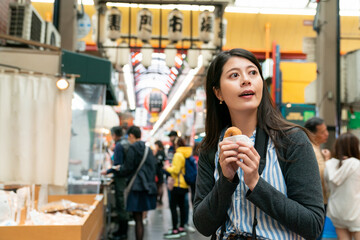 curious asian woman visitor looking at distance while eating donut and visiting kuromon ichiba local market in Osaka japan