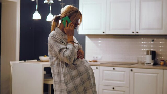 Smiling pregnant female standing in the kitchen and talking on phone with a doctor, consulting online via smartphone. Expectant mother with mobile phone indoors. Happy pregnant woman stroking belly 