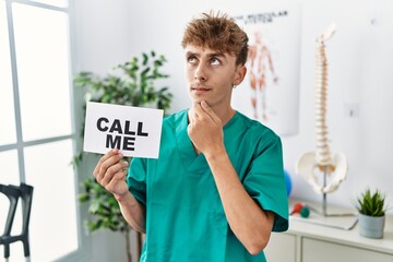 Young caucasian physio man holding call me banner at the clinic serious face thinking about...