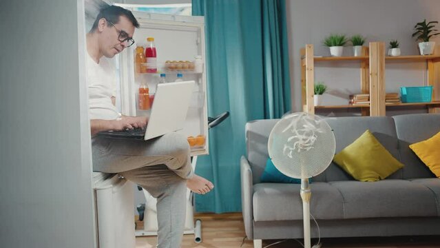freelance man suffering from a heat problem in the living room is sitting in the refrigerator with a laptop
