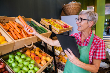Mature woman is working at fruits and vegetables shop She is examining carrot.