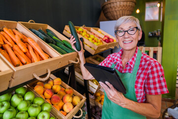 Mature woman is working at fruits and vegetables shop She is examining cucumber.