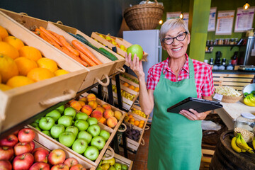 Mature woman is working at fruits and vegetables shop She is examining apples.
