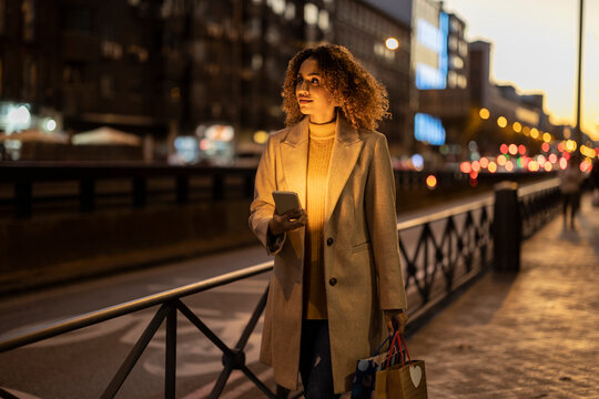 Woman holding shopping bags and mobile phone on footpath at dusk