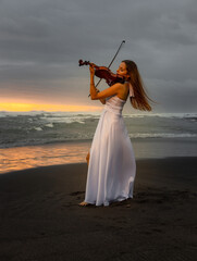 Caucasian woman with violin on the beach. Music and art concept. Slim girl wearing long white dress and playing violin in nature. Sunset time. Cloudy sky. Bali