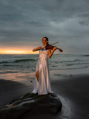Talented woman with violin on the beach. Music and art concept. Slim girl wearing long white dress and playing violin in nature. Sunset time. Cloudy sky. Bali © Olga