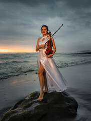 Beautiful Caucasian woman with violin on the beach. Music and art concept. Slim girl wearing long white dress and holding violin in nature. Sunset time. Cloudy sky. Bali