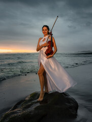 Beautiful Caucasian woman with violin on the beach. Music and art concept. Slim girl wearing long...