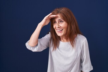 Middle age woman standing over blue background very happy and smiling looking far away with hand over head. searching concept.
