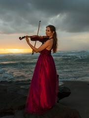 Charming Caucasian woman with violin on the beach. Music and art concept. Slim girl wearing long red dress and playing violin in nature. Sunset time. Cloudy sky. Bali